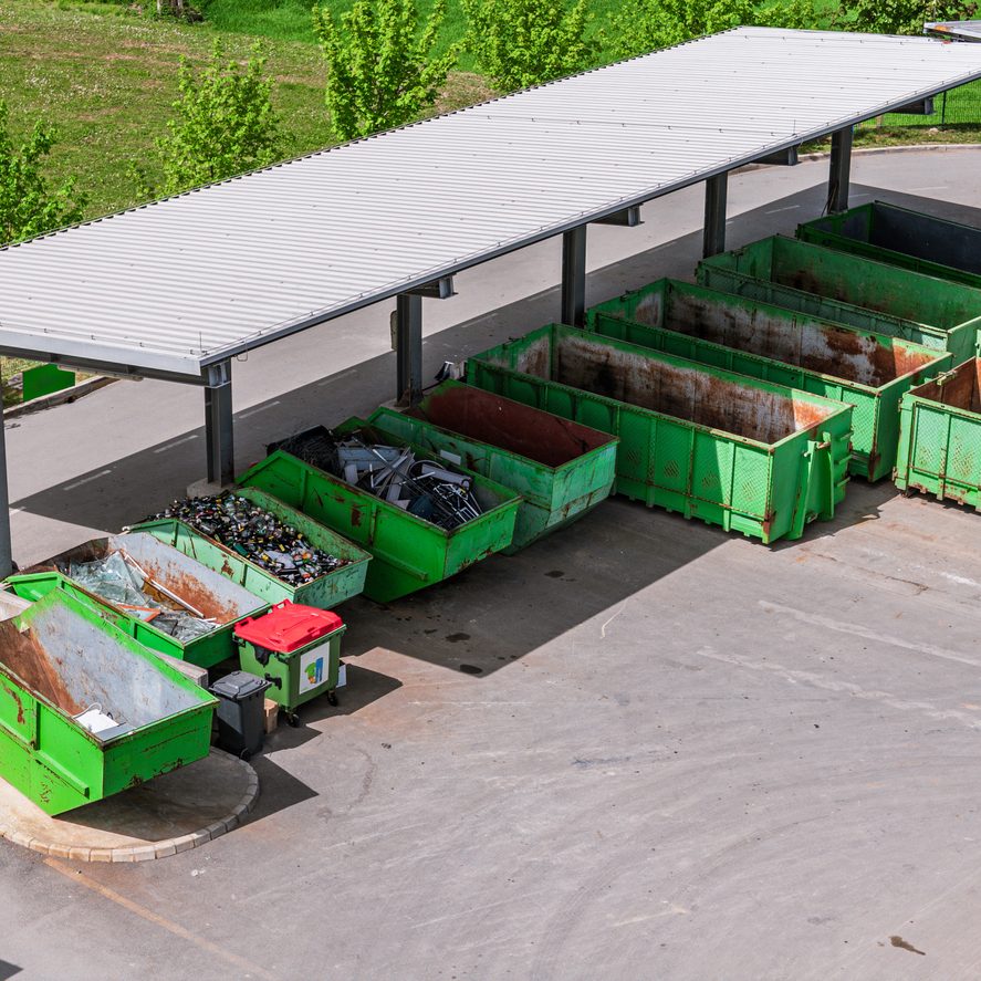 Aerial view of a waste disposal area featuring multiple green dumpsters under a shelter. The site is organized with various containers for recycling and waste collection, surrounded by greenery.