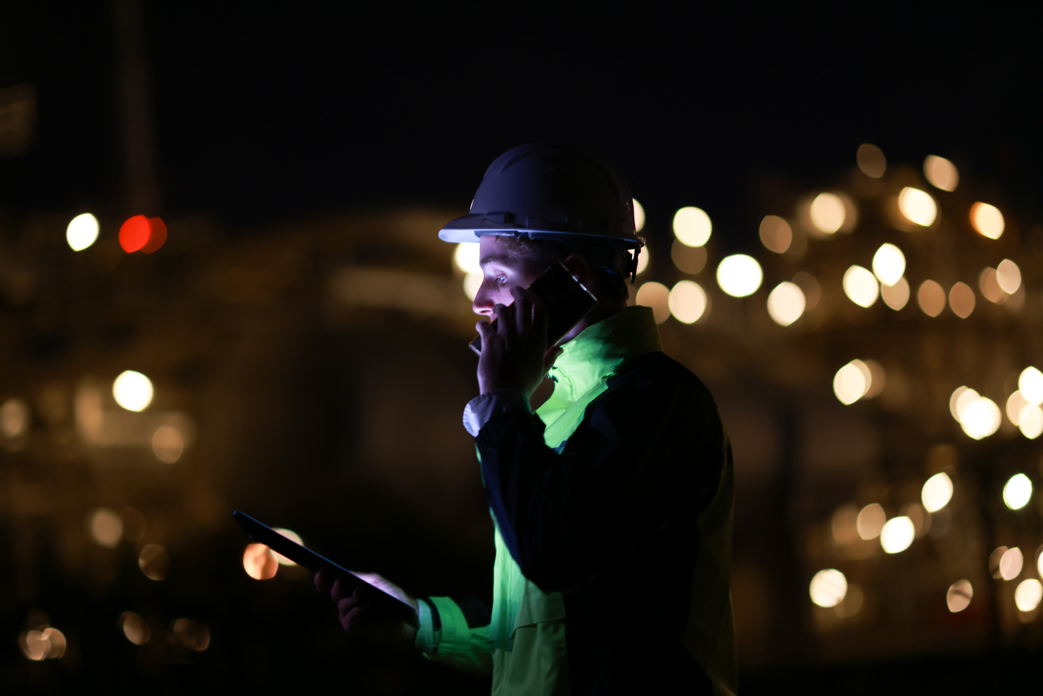 Smart engineer with green safety jacket works on a digital tablet and using a mobile phone at the refinery plant during night shift