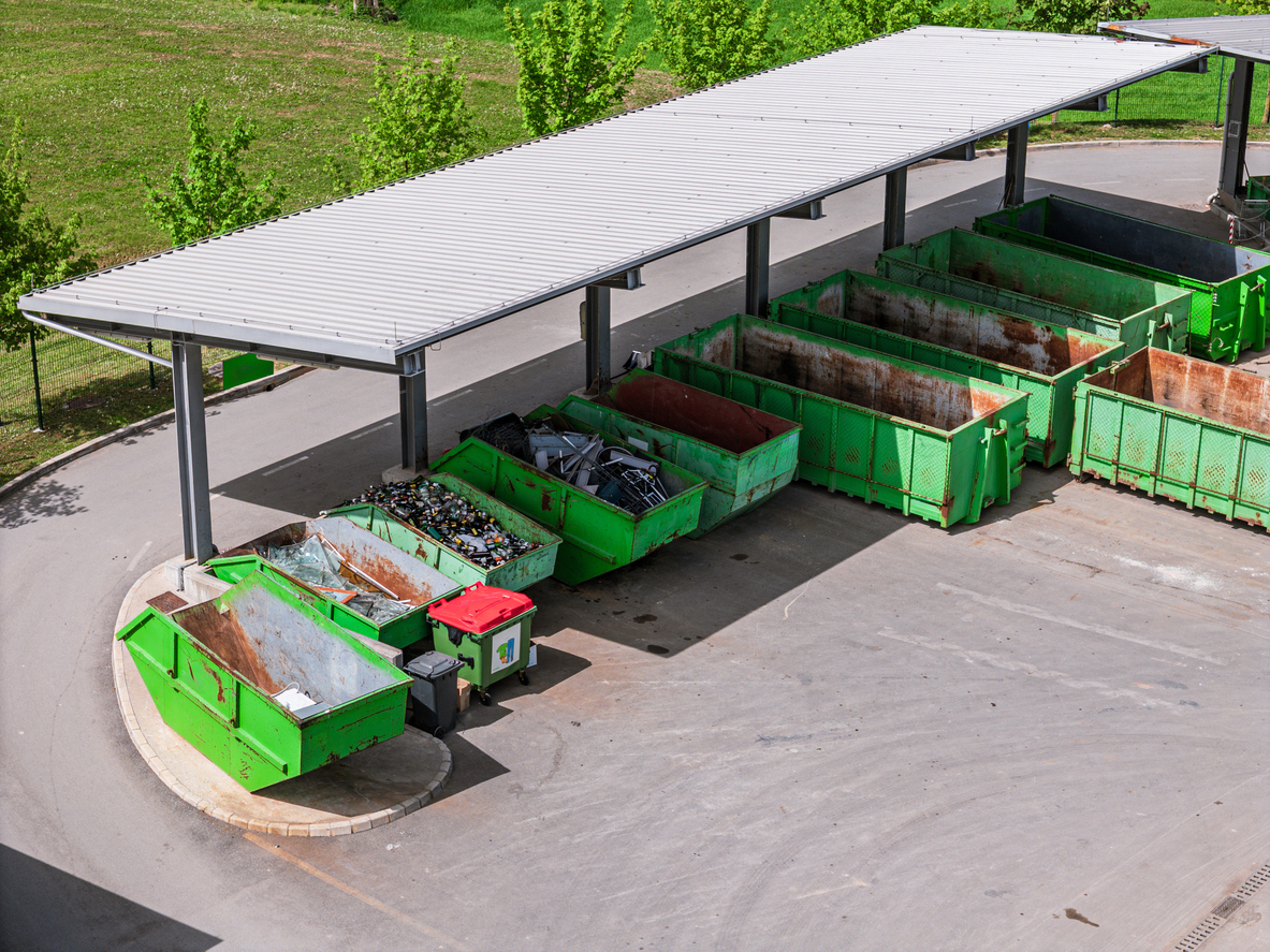 Aerial view of a waste disposal area featuring multiple green dumpsters under a shelter. The site is organized with various containers for recycling and waste collection, surrounded by greenery.